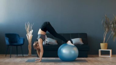 Bir fitness meraklısı şık bir oturma odasında yoga topu üzerinde dengeleyici bir egzersiz yapar. Kurulumda, sağlıklı bir yaşam tarzı ve ev çalışmalarının vurgulandığı modern bir dekor yer alıyor..