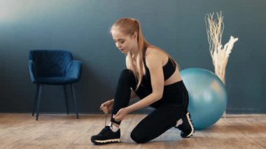 Çağdaş bir spor salonunda ayakkabı bağcıklarını bağlayan genç bir kadın yerde oturuyor. Antrenman kıyafeti giymiş, spor malzemelerinin yanında egzersiz seansına hazırlanıyor..