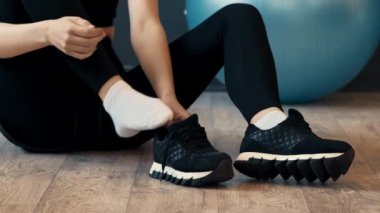 Aktivite kıyafeti giymiş biri spor salonunda oturup spor ayakkabılarını düzeltirken arka planda fitness topu görülebiliyor ve sıradan bir egzersiz ortamı sergiliyor..