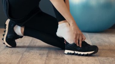 Tahta zeminde diz çöküp ayakkabı bağcıklarını bağlayan bir insan antrenmana hazırlanıyor. Siyah spor ayakkabısı ve tayt giyiyorlar. Yanında da spor topu var..