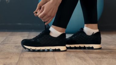 Modern bir spor salonunda spor yapmaya hazırlanan bir insan diz çöker ve ayakkabılarını siyah spor ayakkabılarına bağlar. Çevrede ahşap bir zemin ve yakınında mavi bir egzersiz topu var..