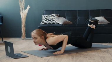 Genç bir kadın, online egzersiz rutinini takip etmek için dizüstü bilgisayar kullanarak ev egzersizi yapıyor. Yoga minderinde egzersiz yaparken formuna odaklanmış..
