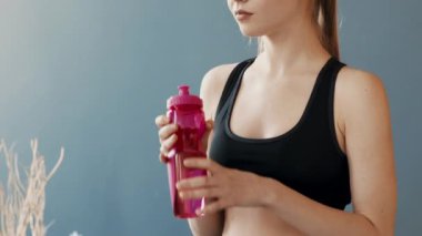 Siyah kolsuz tişörtlü genç bir kadın pembe bir su şişesinden su içiyor. Kapalı alanda yapılan egzersizden sonra su kaybediyor. Ayarlar yumuşak bir arkaplan ve onun faaliyetlerine odaklanmayı içeriyor.