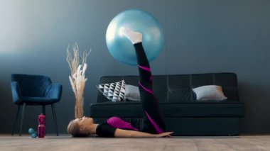 Aktif giyimli bir kadın, çağdaş bir yaşam alanında dengeli bir top kullanarak çekirdek gücü ve uygunluğuna odaklanmış bir egzersiz yapıyor. Odada minimalist dekor ve yeterli ışık var..