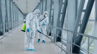 İki işçi, modern koridor boyunca dezenfektan püskürtüyor. Çevre parlak ve ferah, sağlık krizlerinde hijyenin önemini vurguluyor..