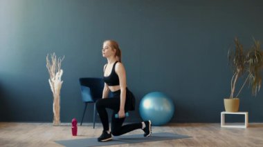 Genç bir kadın, bitki ve spor malzemeleriyle süslenmiş, geniş, parlak ışıklı bir spor salonunda dambıllarla hareket egzersizi yapıyor..