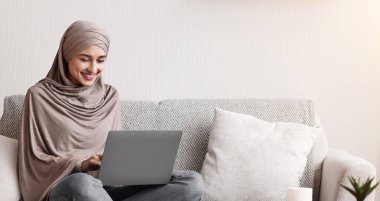 Modern Arap kız evde dizüstü bilgisayar kullanıyor, oturma odasında kanepede otururken uzaktan çalışıyor.