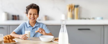 Genç siyah bir çocuk mutfak masasında oturuyor, elinde kurabiye ve bir bardak süt tutarken gülümsüyor. Konforlu ortamda bir kase kurabiye ve bir sürahi süt var. Öğleden sonra ziyafeti için mükemmel..