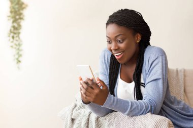 Heyecanlı siyahi kadın internetten iyi haberler alıyor, evdeki koltukta oturuyor, boş bir alanda.