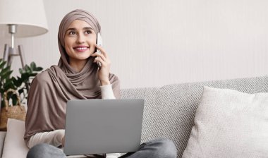 Modern İletişim. Evde dizüstü bilgisayar kullanırken cep telefonuyla konuşan başörtülü neşeli Arap kız rahat koltukta oturuyor, boş yer var.