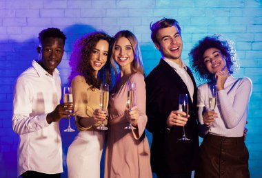 Şampanya bardaklı mutiryaki gençler. Kapalı alanda duvarın karşısında duran şirket etkinliğini kutlayan kameraya gülümsüyorlar.