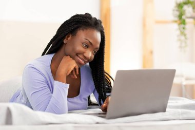 Laptop kullanan gülümseyen Afro Öğrenci Kız. Yatak odasında yatarken video görüşmesi yapıyor. Haftasonu Sabah Kavramı. Seçici Odaklanma