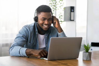 Neşeli Afro-Amerikan işçisi kulaklık takıyor, ofiste dizüstü bilgisayarda webinar izliyor.