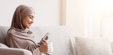 Haftasonu rahatla. Başörtülü Müslüman kız evde akıllı telefon ve kahveyle dinleniyor, oturma odasında koltukta oturuyor, boş yer var.