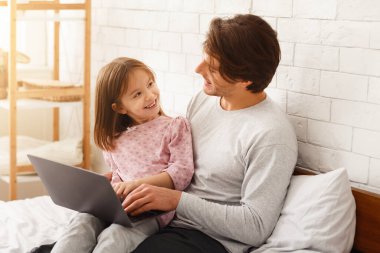 Küçük kız ve babası yatak odasında dizüstü bilgisayar kullanıyorlar, hafta sonlarını birlikte planlıyorlar, boşluğu kopyalıyorlar.