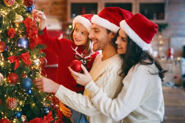 Kırmızı Noel Baba şapkalı sevimli aile evde baloncuklar ve ışıklarla Noel ağacı süslüyorlar.