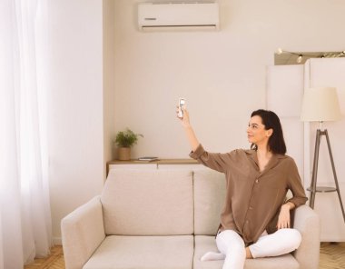 Ev Hayatı, İklim Kontrolü. Kanepede otururken klimayı açan mutlu bir kadın.