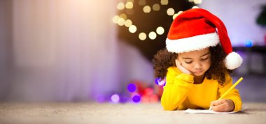 Konsantre olmuş küçük afro kız Noel Baba 'ya mektup yazıyor, Noel ağacının yanında yerde yatıyor, boş yer var.