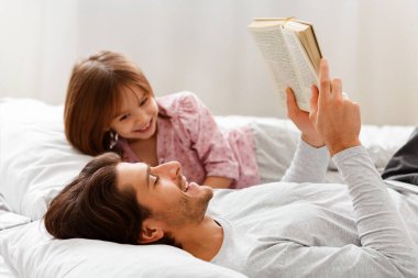 Genç baba, neşeli kızına kitap okuyor, birlikte yatağa uzanıyor.