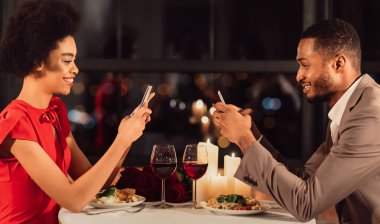 Modern Relationship. Happy Afro Couple Using Smartphones During Romantic Date In Restaurant. Panorama