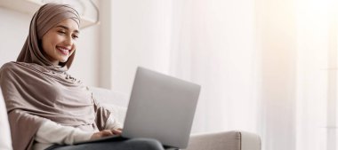 Serbest Kariyer Konsepti. Başörtülü genç Arap kadın evde dizüstü bilgisayarla çalışıyor bilgisayar ekranına bakıyor ve gülümsüyor.