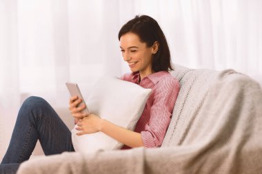 Boş zaman. Yastık tutan mutlu genç bir kadın, koltukta oturuyor, akıllı telefon kullanıyor, kopyasal alan kullanıyor.