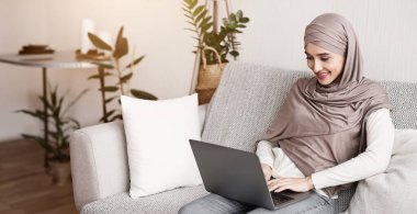 Başörtülü genç Müslüman kadın evde dizüstü bilgisayar kullanıyor, klavyede yazıyor, e-postaları cevaplıyor, arkadaşlarıyla çevrimiçi sohbet ediyor..