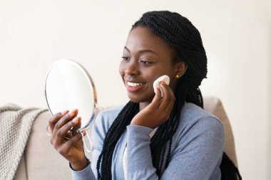 Günlük Skincare Rutini. Gülümseyen siyah kadın aynaya bakıyor ve pamuklu bezle yüzünü temizliyor, evdeki kanepede oturuyor, boş yer var.