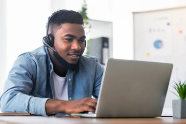 İş yerindeki çağrı merkezi operatörü. Yakışıklı siyah yönetici kulaklık takıyor ve dizüstü bilgisayarda daktilo yazıyor, modern ofiste çalışıyor, boş yer var.