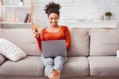 İnternetten alışveriş. Laptop ve kredi kartıyla gülümseyen Afro kadın evdeki koltukta oturmuş internetten bir şeyler alıyor.