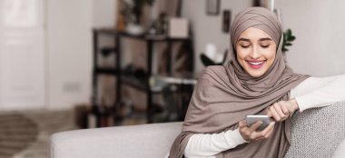 Tesettürlü neşeli Müslüman kadın evde akıllı telefon kullanıyor, internette geziniyor ya da sosyal ağda geziniyor, cihaz ekranına bakıyor ve gülümsüyor.