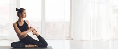 Genç bir kadın yoga pozu veriyor, geniş pencereli iyi aydınlatılmış bir stüdyoya odaklanıyor, boşluğu kopyalıyor.