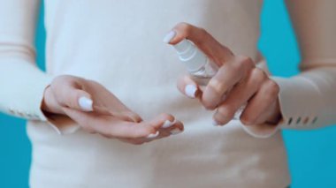 Bir kişi içeride dururken avuç içine el dezenfektanı koyuyor. Canlı mavi arka plan, açık renkli kazakları ve manikürlü tırnaklarıyla çelişiyor..