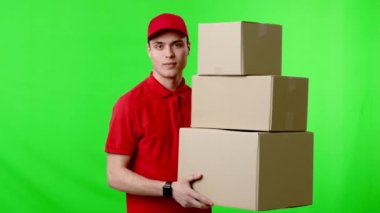 Kırmızı üniformalı ve şapkalı bir teslimatçı, nakliyeye ya da dağıtıma hazır, canlı yeşil bir zemin üzerinde üç karton kutu taşırken gülümsüyor..