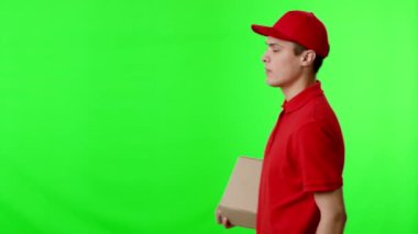 Kırmızı tişörtlü ve şapkalı bir teslimatçı elinde küçük bir paket tutarken gülümsüyor, canlı yeşil bir arkaplanın önünde duruyor, genellikle dijital düzenleme için kullanılır..