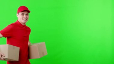 Kırmızı şapkalı ve gömlekli neşeli bir teslimatçı, bir sonraki teslimat için canlı yeşil bir arkaplanın önünde dururken gülümseyen iki karton kutu taşıyor..