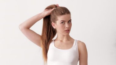 Genç bir kadın, uzun saçlarını at kuyruğu şeklinde şekillendirirken minimalist bir alanda duruyor. İzleyiciye kendinden emin bakıyor, zarif ve sıradan bir saç stili sergiliyor..