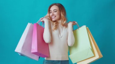 Neşeli genç bir kadın elinde çeşitli renkli alışveriş çantaları tutarken gülümsüyor. Alışveriş deneyiminin tadını çıkarıyor, canlı mavi bir arkaplan önünde mutluluk saçıyor..