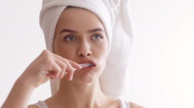Dişlerini fırçalamaya odaklanmış, iyi ışıklandırılmış bir banyoda duran bir kadın. Kafasına havlu takıyor ve sabah hijyen rutinini uygularken mutlu görünüyor..