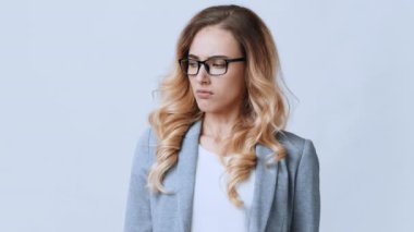 Dalgalı sarı saçlı ve gözlüklü genç bir kadın gri bir ceket giyer, düşünceli bir şekilde gözlerini kaçırır ve minimalist bir ortamda kendine güvenir..