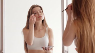 Genç bir kadın iyi ışıklandırılmış bir yerde duruyor, aynaya bakarken nazikçe göz farı sürüyor. Uzun saçları var ve basit bir bluz giyiyor. Makyaj rutinine odaklanıyor..