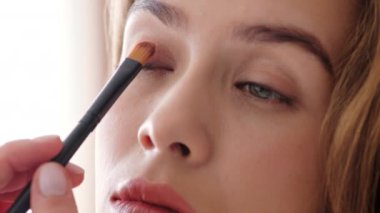 Bir insan, küçük bir fırçayla göz kapağına nazikçe göz makyajı yapar. Ayarlar, makyaj uygulama sürecine odaklanmayı artıran yumuşak ve doğal ışıklandırmayı içeriyor.