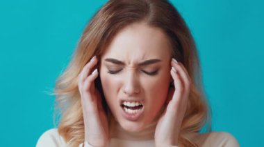 Uzun, dalgalı saçlı genç bir kadın kafasını tutarak rahatsızlığını ifade eder. Parlak mavi arka plan onun gergin yüz ifadesini vurguluyor, stres ve hayal kırıklığını vurguluyor..