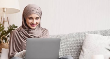 Booking vacation online. Portrait of young muslim woman in hijab using laptop, making hotel and tickets reservation while sitting on couch at home