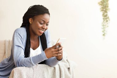 Gülümseyen Afrikalı Amerikalı kadın evinde akıllı telefon kullanıyor. Kanepede dinlenirken mesajlaşıyor ya da sosyal ağlara bakıyor.