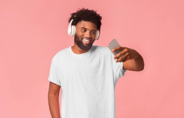Kablosuz kulaklıklı neşeli afro kız pembe arka planda telefonla selfie çekiyor.