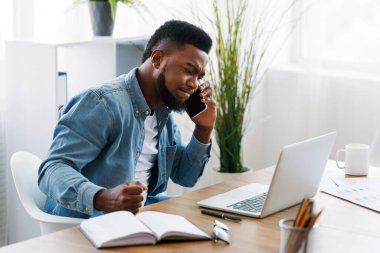 İşle ilgili sorunlar. Stresli Afro-Amerikan iş adamı telefonda konuşuyor, dizüstü bilgisayara bakıyor ve hayal kırıklığı içinde yumruğunu sıkıyor..