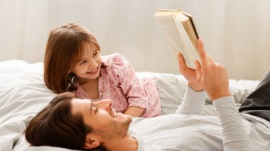Genç adam, küçük gülümseyen kızına kitap okuyor, birlikte yatağa uzanıyor.
