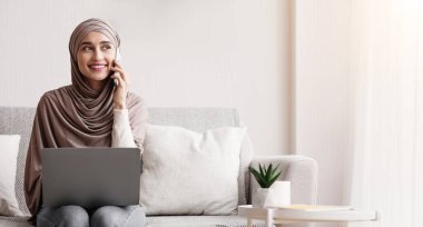 Evde dizüstü bilgisayar kullanırken cep telefonuyla konuşan tesettürlü gülümseyen İslamcı kız, boş alan.