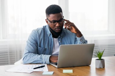 Endişeli Afro-Amerikan çalışanı cep telefonuyla konuşuyor ve laptop ekranına bakıyor, projeyle ilgili sorunları var, boşluğu kopyala